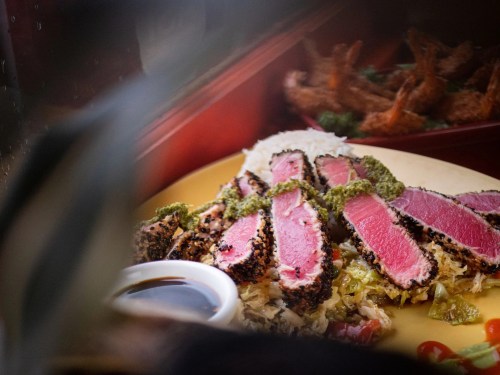 A plate of sliced seared tuna with a green sauce, served with vegetables and a small dish of soy sauce.
