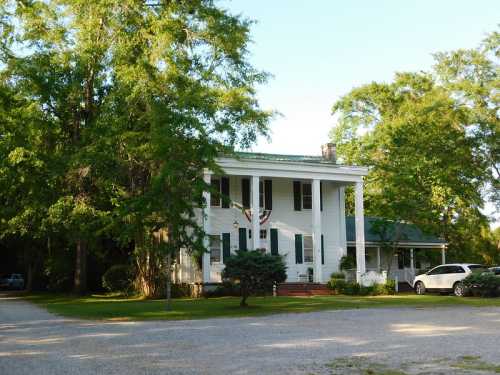 A white two-story house with columns, surrounded by trees and a gravel driveway. Flags hang from the porch.