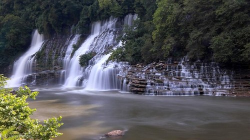 A serene waterfall cascading over rocky cliffs, surrounded by lush green trees and a calm river below.