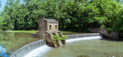 A stone structure sits on a small dam, with water cascading over it, surrounded by lush green trees.