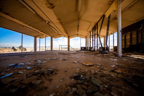 Abandoned interior of a building with broken glass on the floor and large windows revealing a desert landscape outside.
