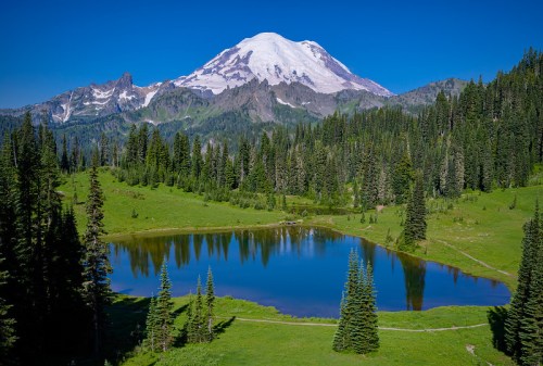 A serene landscape featuring a clear lake reflecting a snow-capped mountain, surrounded by lush green forests and blue skies.