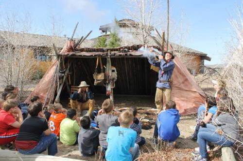 A group of children watches a demonstration outside a traditional shelter, with one person showcasing a skill.