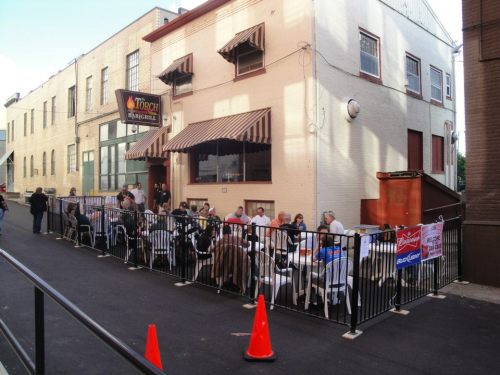 Outdoor dining scene with a long table set up along a building, surrounded by people enjoying a meal.