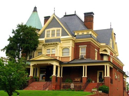A large, historic Victorian-style house with a steep roof, ornate details, and a green turret, surrounded by greenery.