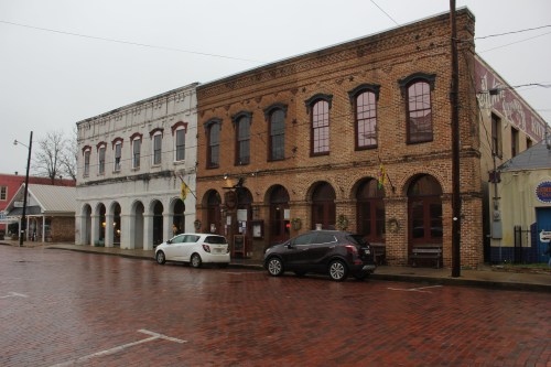 Historic brick buildings line a wet street, with parked cars and flags hanging from the windows on a cloudy day.