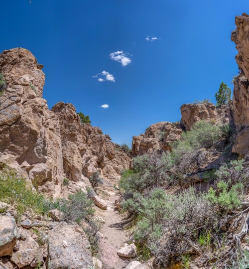 A narrow canyon surrounded by tall rock formations and sparse vegetation under a clear blue sky with a few clouds.