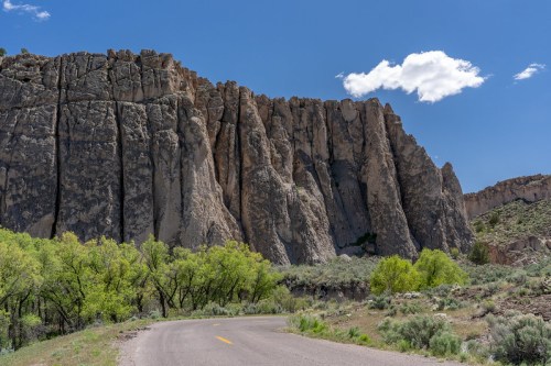 A winding road curves around towering rock formations under a blue sky with a few clouds. Lush greenery lines the road.