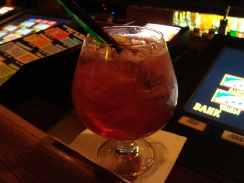 A close-up of a cocktail in a round glass with ice, sitting on a bar counter with gaming machines in the background.