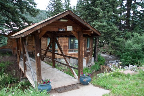 A wooden cabin with a bridge leading to it, surrounded by trees and greenery, near a flowing stream.