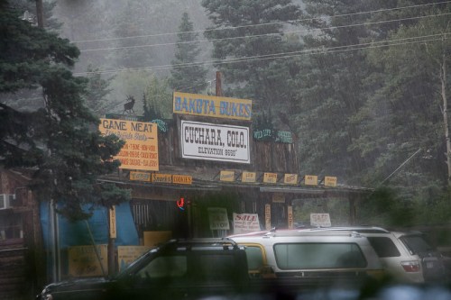 A rustic store sign reading "Dakota Buck's" in Cuchara, Colorado, amidst heavy rain and surrounded by trees.