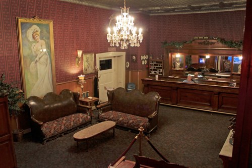 A vintage lobby with ornate wallpaper, a chandelier, plush sofas, and a large painting of a woman on the wall.