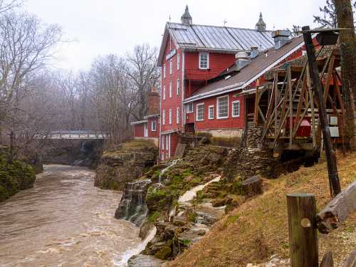 A red historic building beside a rushing river, surrounded by bare trees and rocky terrain on a cloudy day.