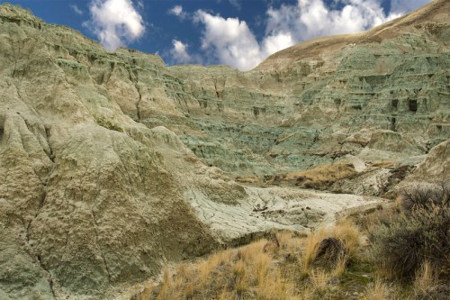 A rugged landscape of greenish rock formations and dry grass under a partly cloudy sky.