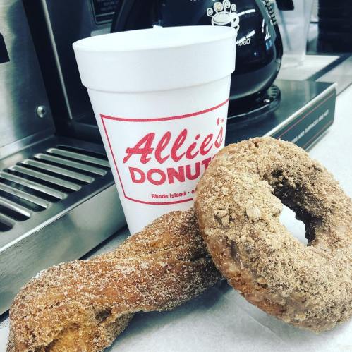 A cup of coffee from Allie's Donuts next to two freshly baked donuts on a countertop.