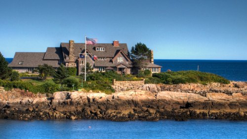 A coastal mansion with a flag, surrounded by greenery and rocky shoreline, overlooking a calm blue ocean.