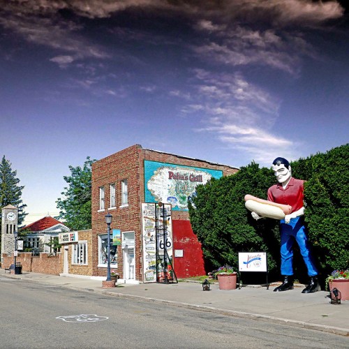 A colorful roadside scene featuring a large statue of a man holding a hot dog, with vintage buildings and greenery.