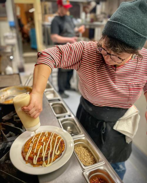A chef in a striped shirt and beanie drizzles sauce on a plate of food in a busy kitchen.