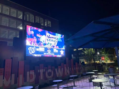 A large outdoor screen displays a baseball game at night, with fans holding signs in a lively urban setting.