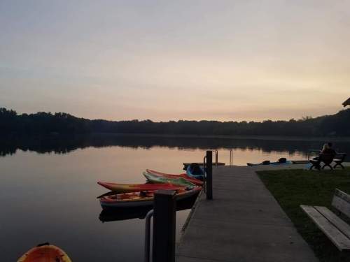 A serene lake at sunset with colorful kayaks lined up on the shore and a person sitting on a bench nearby.