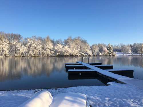 A serene winter scene featuring a snowy landscape, calm water, and a wooden dock extending into the lake.