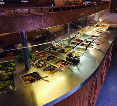 A buffet bar with various food items in containers, including salads, toppings, and dressings, under a glass shield.