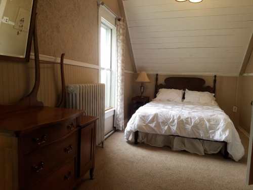 Cozy bedroom with a sloped ceiling, featuring a bed, dresser, lamp, and a window with curtains.