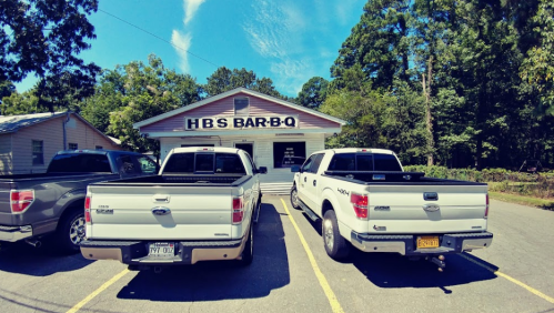 Two white pickup trucks parked in front of a restaurant with "HBS BAR-B-Q" sign, surrounded by trees and blue sky.