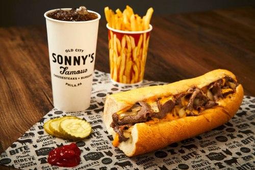 A cheesesteak sandwich with fries and a drink on a patterned paper, featuring pickles and ketchup on the side.