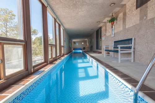 Indoor swimming pool with large windows, blue water, and a bench along the side, surrounded by a light-colored wall.