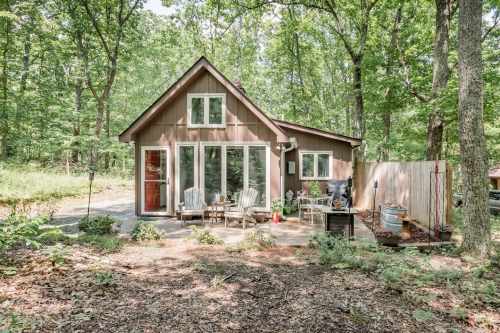 A cozy brown cabin surrounded by trees, featuring a patio with chairs and a grill in a serene forest setting.