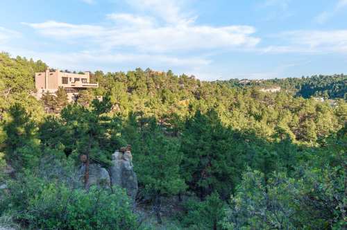 A modern house nestled among lush green trees and rocky formations under a clear blue sky.