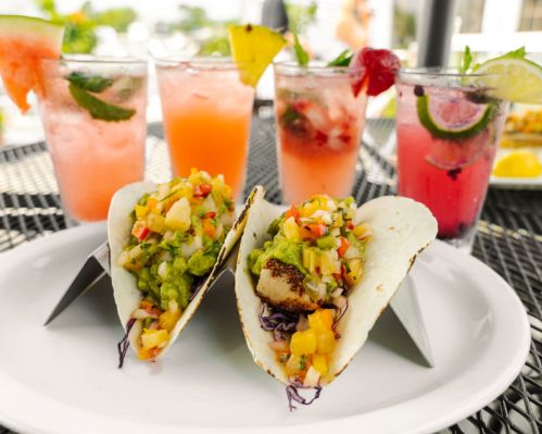 Two fish tacos topped with guacamole and salsa, served on a plate with colorful cocktails in the background.