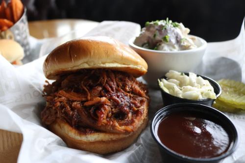 A pulled pork sandwich on a bun, served with coleslaw, potato salad, pickles, and barbecue sauce on the side.