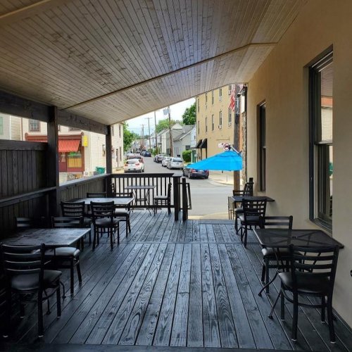 Outdoor seating area with black tables and chairs, overlooking a street with parked cars and a blue umbrella in the distance.