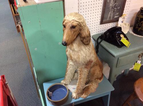 A life-sized wooden sculpture of a seated dog next to a blue bowl, displayed in a vintage shop.