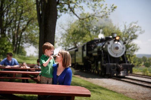 A woman and a young boy share a moment at a picnic table, with a steam train in the background on a sunny day.