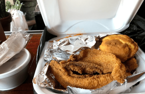 A takeout container with fried fish, a biscuit, and a cup of sauce on a wooden table.