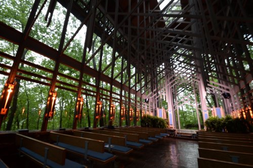 Interior of a modern chapel with wooden beams, surrounded by greenery and soft lighting, creating a serene atmosphere.