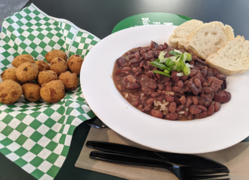 A bowl of red beans and rice topped with green onions, served with cornbread balls and slices of bread on a checkered napkin.