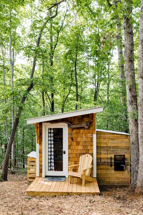 A small wooden cabin with a porch and an Adirondack chair, surrounded by tall trees in a forested area.