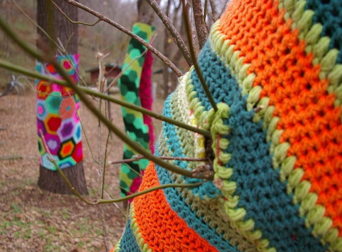 Colorful knitted patterns wrap around tree trunks in a forest setting, with vibrant hues of orange, green, and pink.