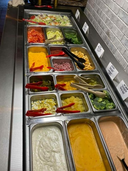 A salad bar with various toppings, including vegetables, cheeses, and dressings, in metal containers.