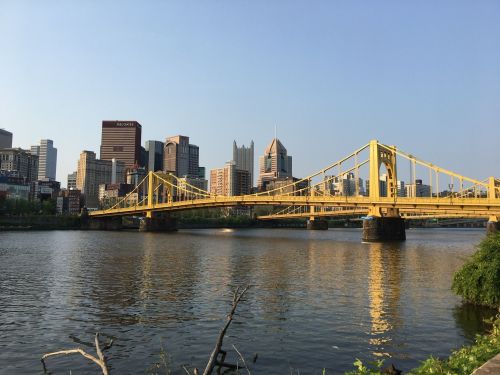 A yellow bridge spans a river with a city skyline in the background under a clear blue sky.