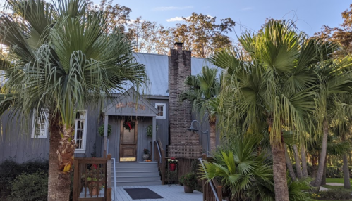 A charming gray house with a chimney, surrounded by lush palm trees and greenery, under a clear blue sky.