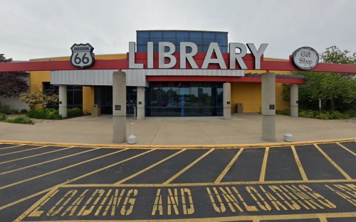 Front view of a modern library building with a large sign, featuring a gift shop and a parking area.