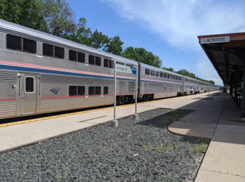 A train at a station with a sign reading "Lacrosse," surrounded by trees and a clear blue sky.