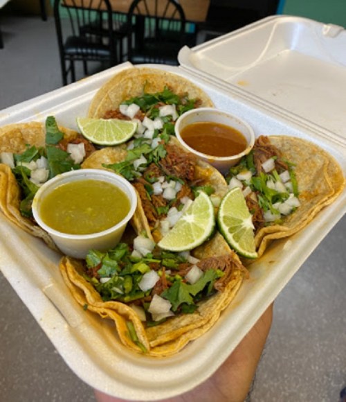 A hand holding a takeout container with four tacos topped with cilantro, onions, lime wedges, and two sauces.
