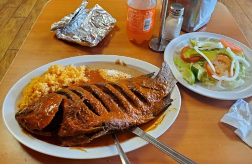 A plate of grilled fish in sauce, served with rice, beans, and a side salad on a wooden table.