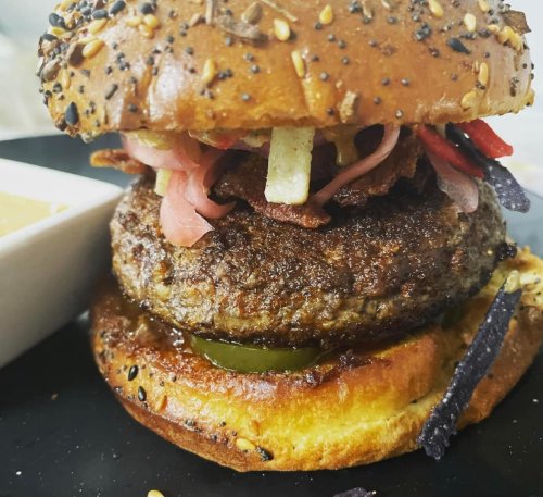 Close-up of a juicy burger with a sesame seed bun, topped with bacon, pickles, and colorful toppings, served with dipping sauce.